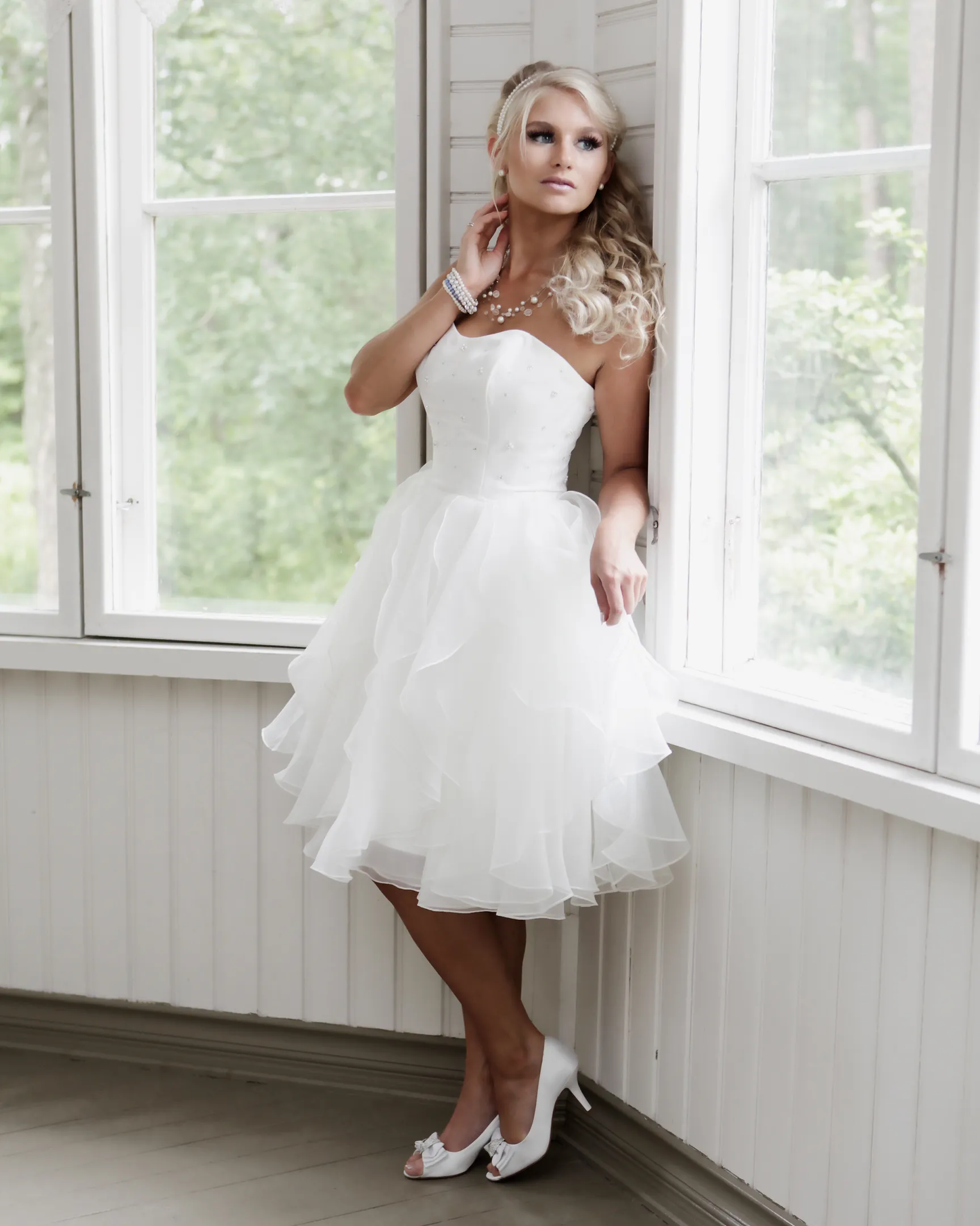 Ruth in white cocktail dress by window in bright veranda