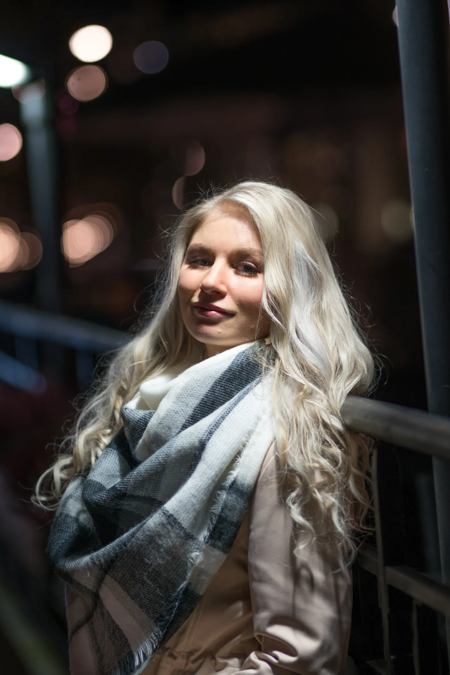 Ruth with plaid scarf leaning on railing, night city bokeh