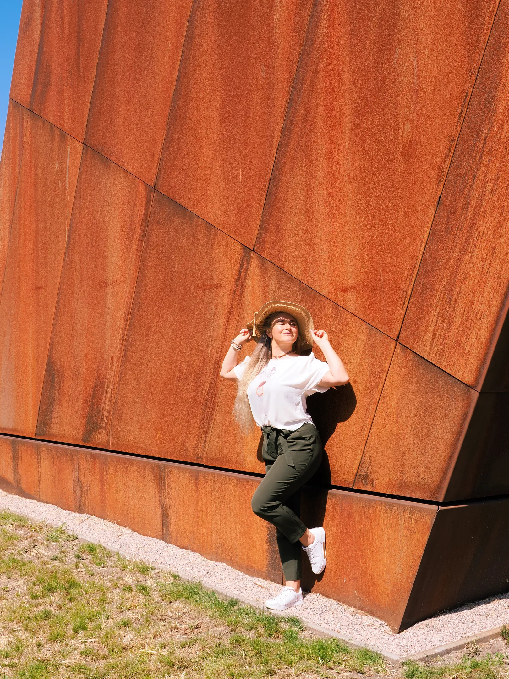 Ruth posing with straw hat against rust-colored wall in sunshine