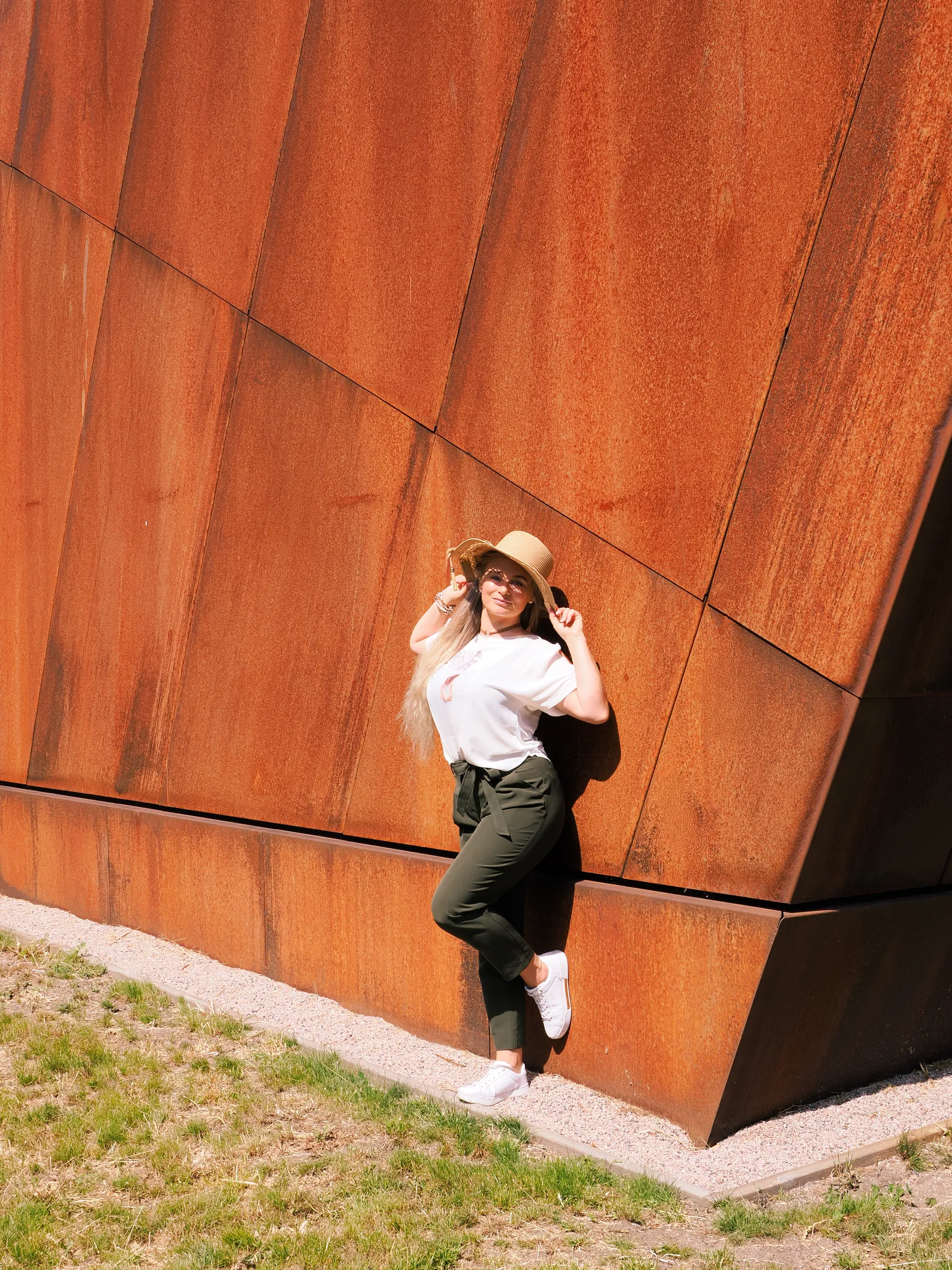 Ruth leaning against rust wall with hat, summery casual pose