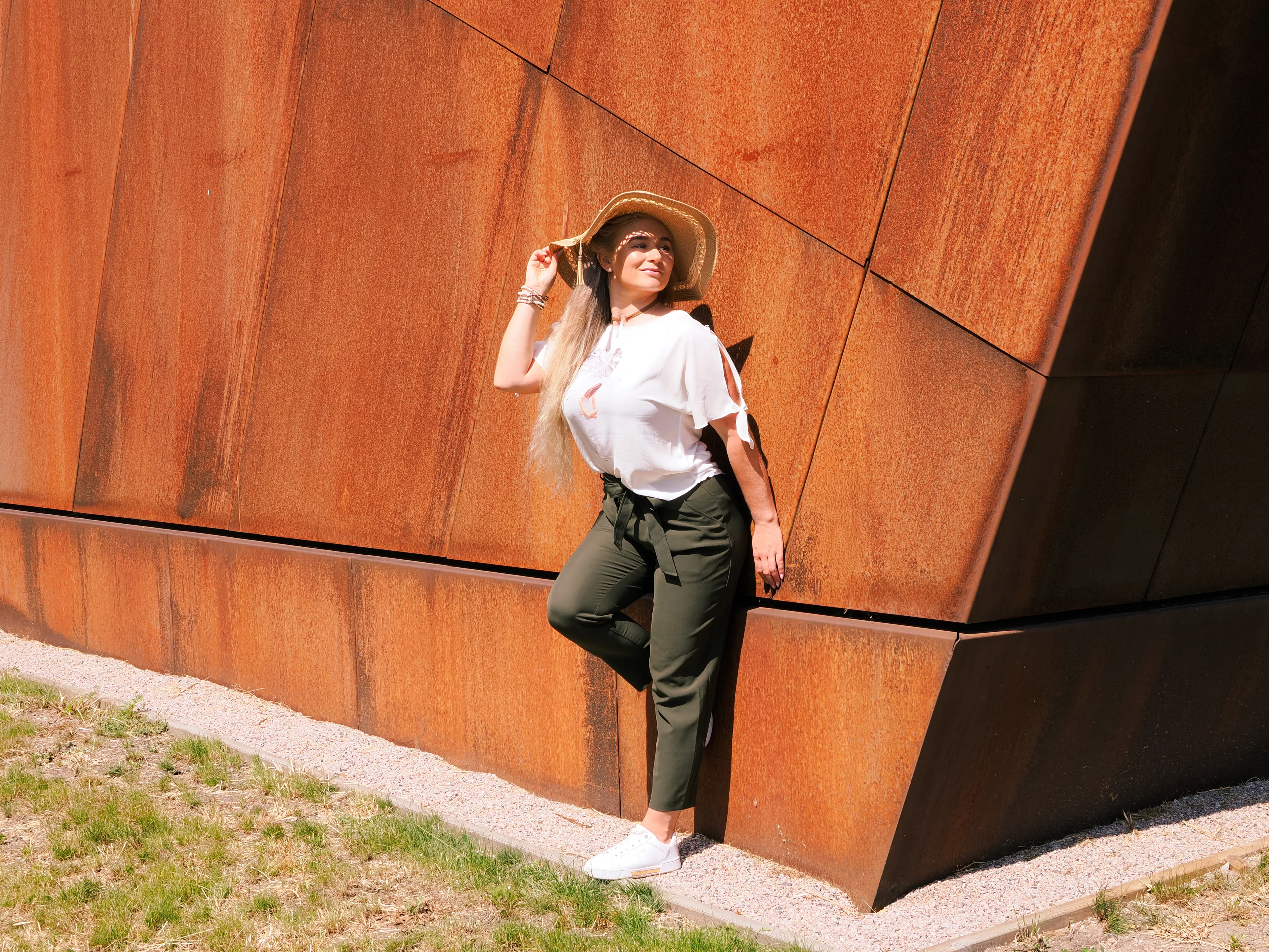 Ruth in white top and hat leaning against corten steel wall