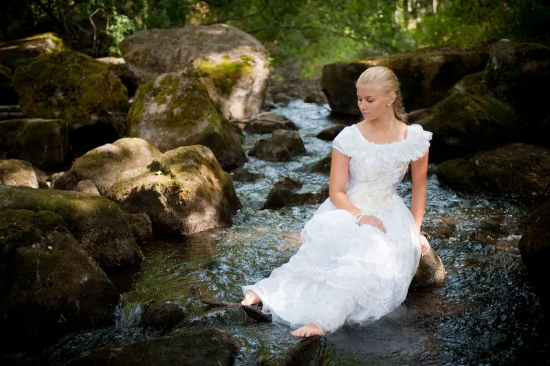 Ruth in white gown sitting in stream among mossy boulders