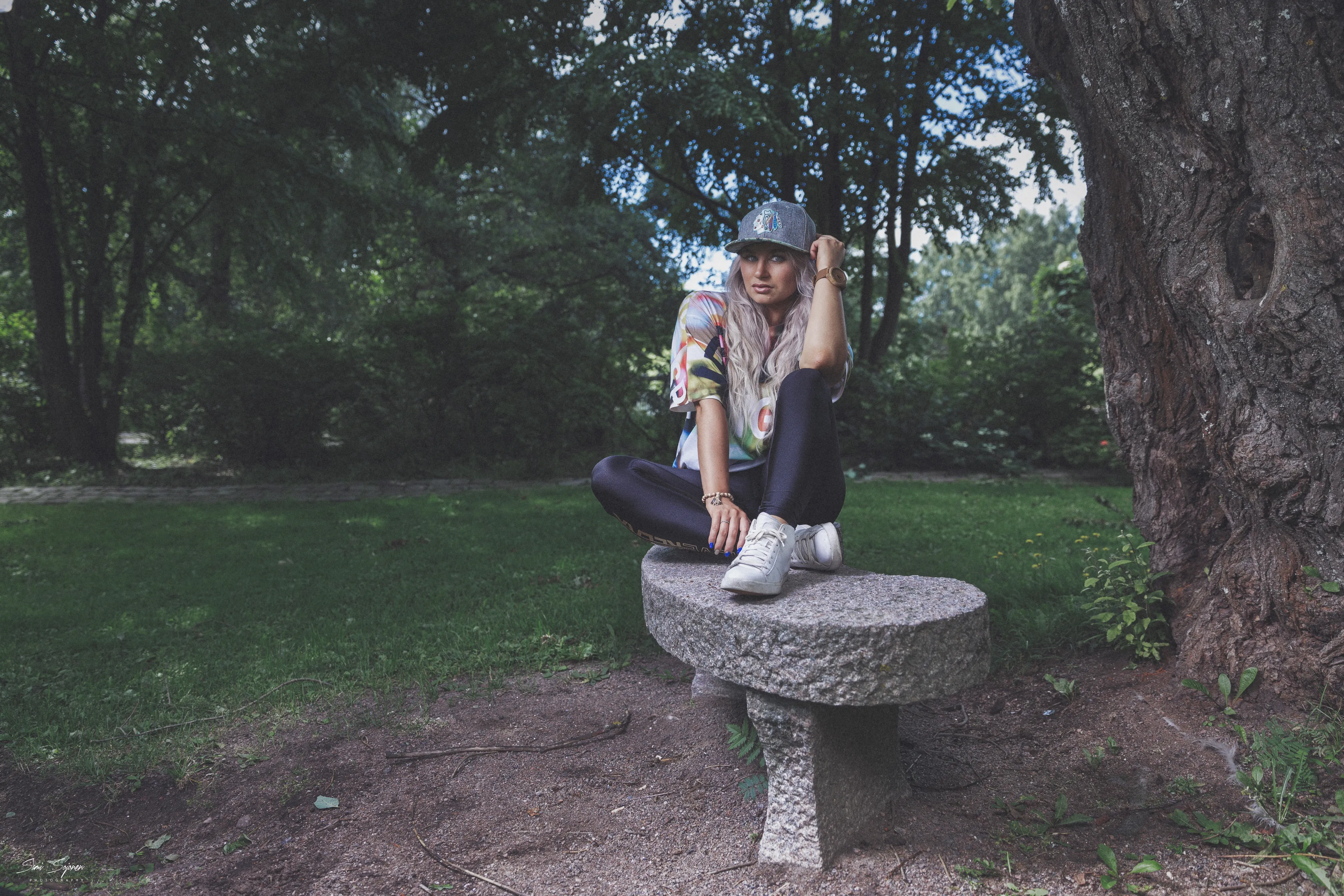 Ruth in colorful top and cap seated cross-legged on park bench