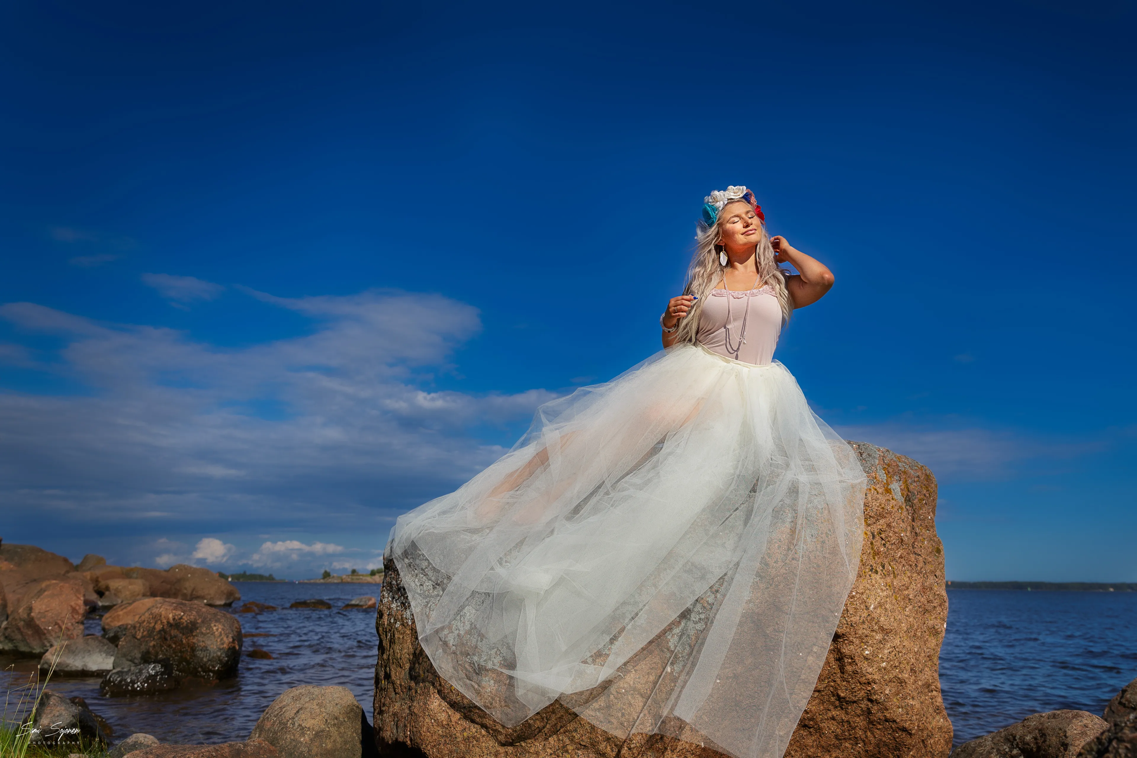 Ruth in tulle gown on seaside boulder under blue sky, ethereal