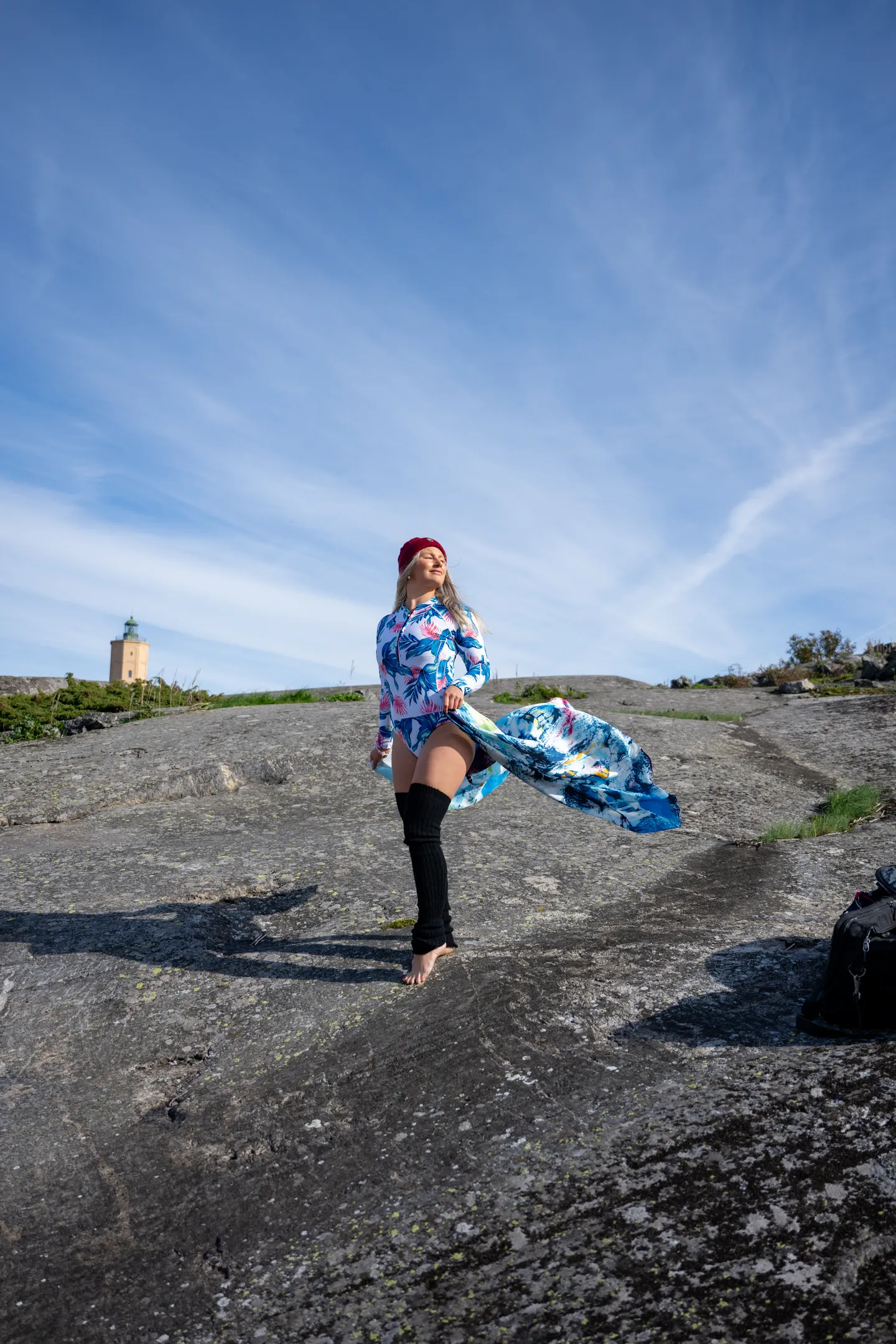 Ruth on rocky coastline in flowing blue-print dress with red beret